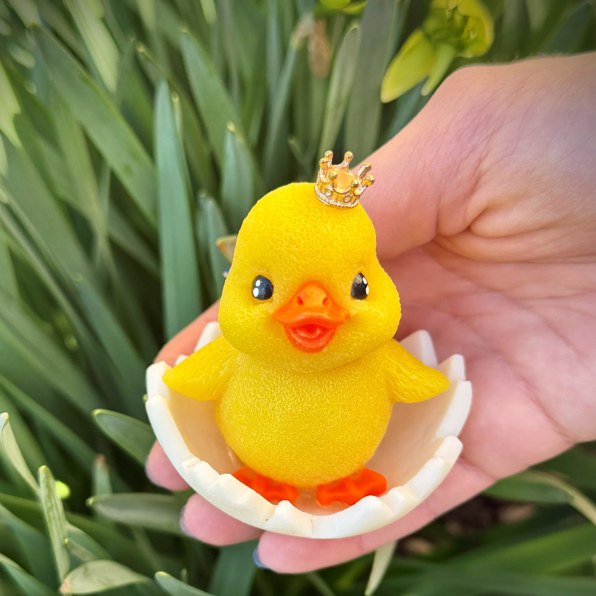 Hand holding a bright yellow chick-shaped handmade soap sitting in a cracked white eggshell design outdoors among green leaves.