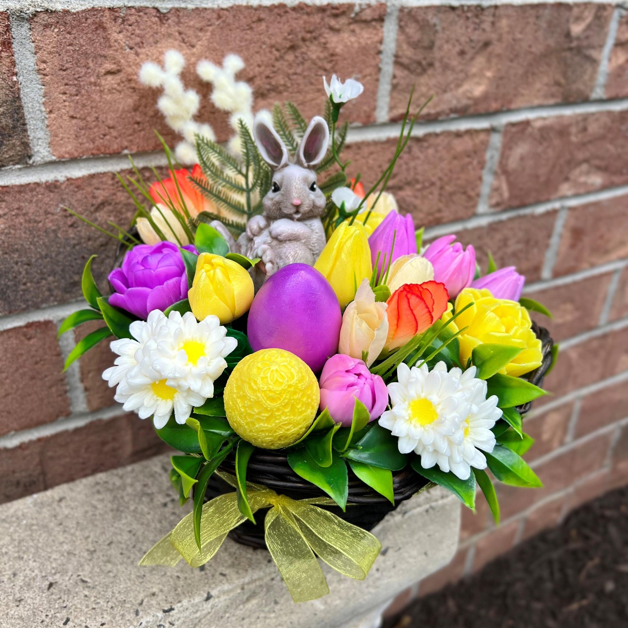 Colorful Easter arrangement with flowers, eggs, and a rabbit figurine against a brick wall.
