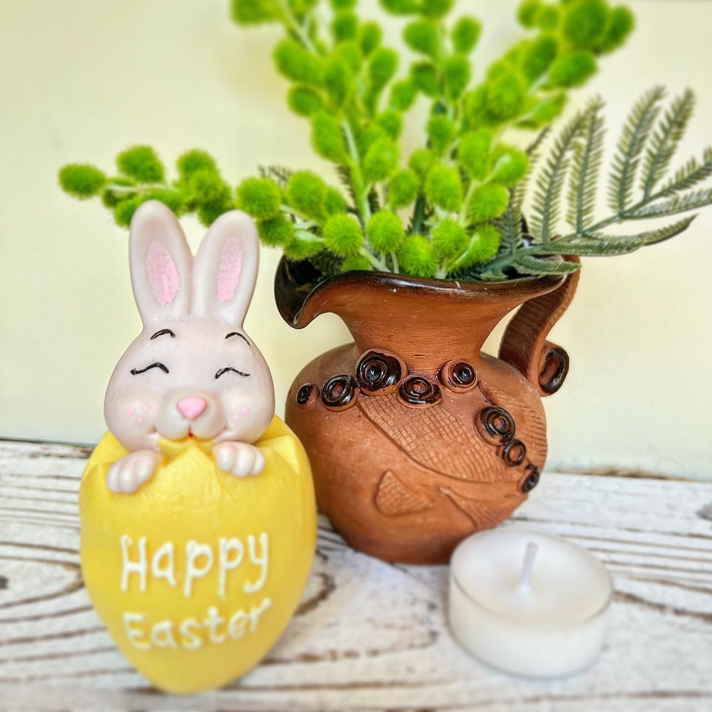 Decorative Easter egg with a bunny and 'Happy Easter' text, next to a brown vase with green plants on a wooden surface.