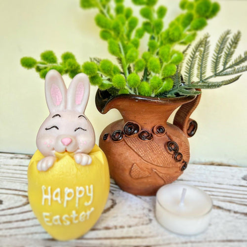 Decorative Easter egg with a bunny and 'Happy Easter' text, next to a brown vase with green plants on a wooden surface.