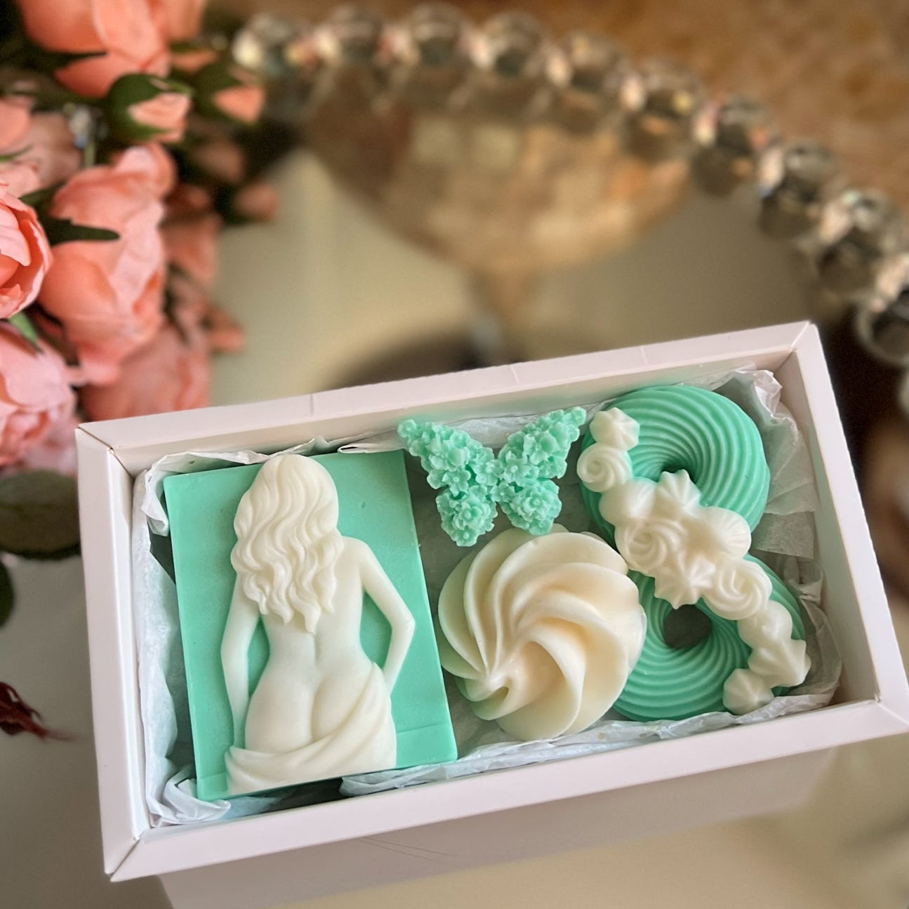 Decorative soaps in a box with a butterfly and floral designs, placed on a reflective surface with flowers in the background.