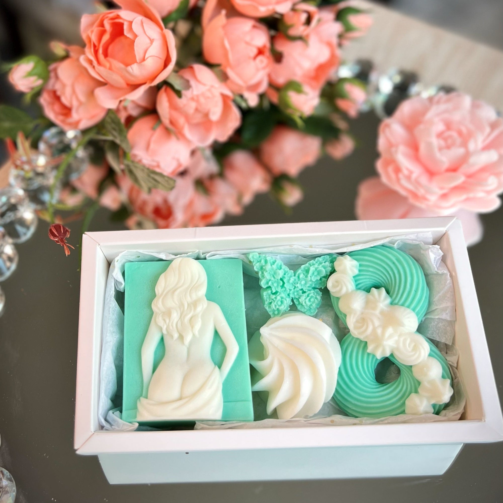 Decorative soap set with figurines and designs in a box, with pink flowers in the background.
