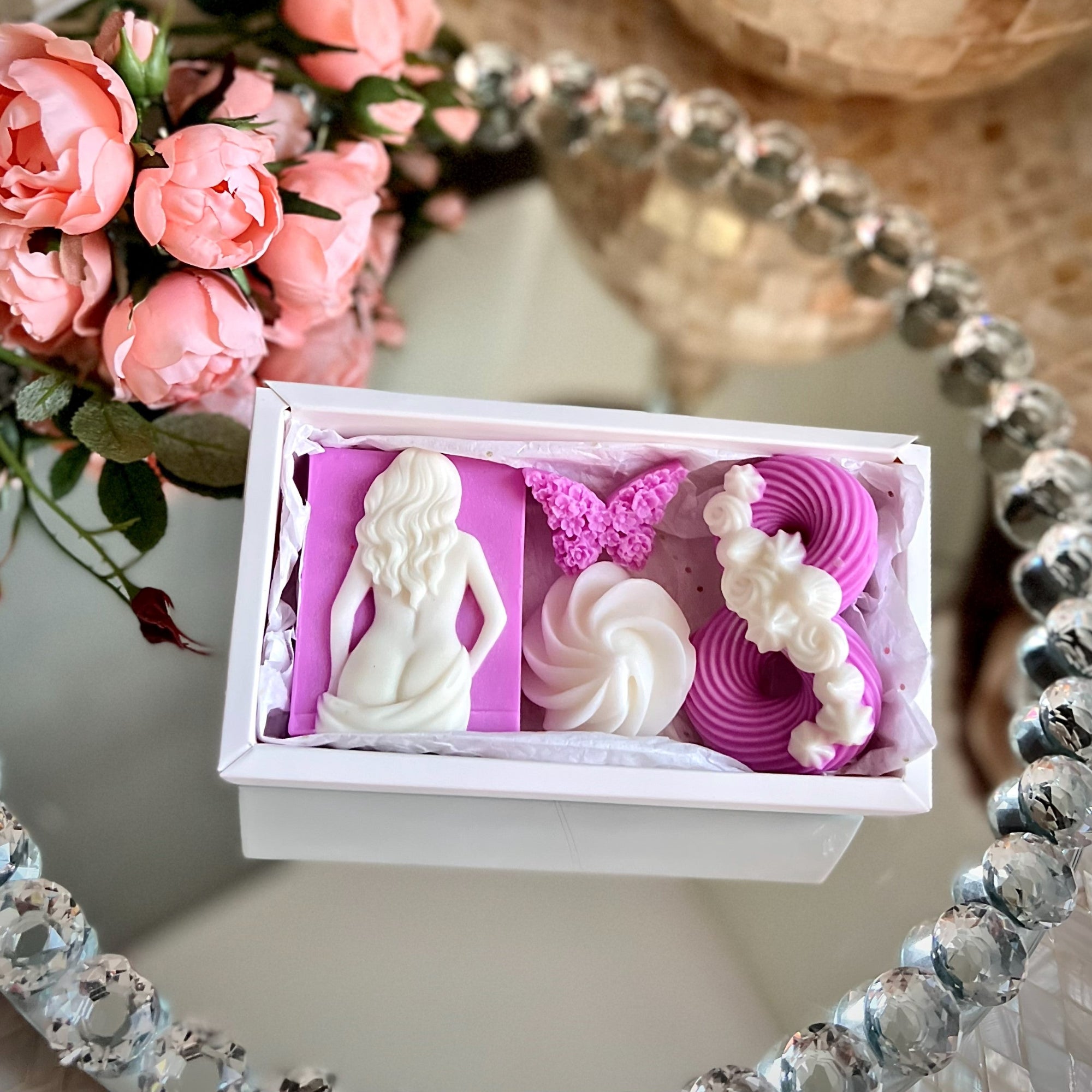 Decorative box with figurines, flowers, and a butterfly on a reflective surface with a floral arrangement in the background.