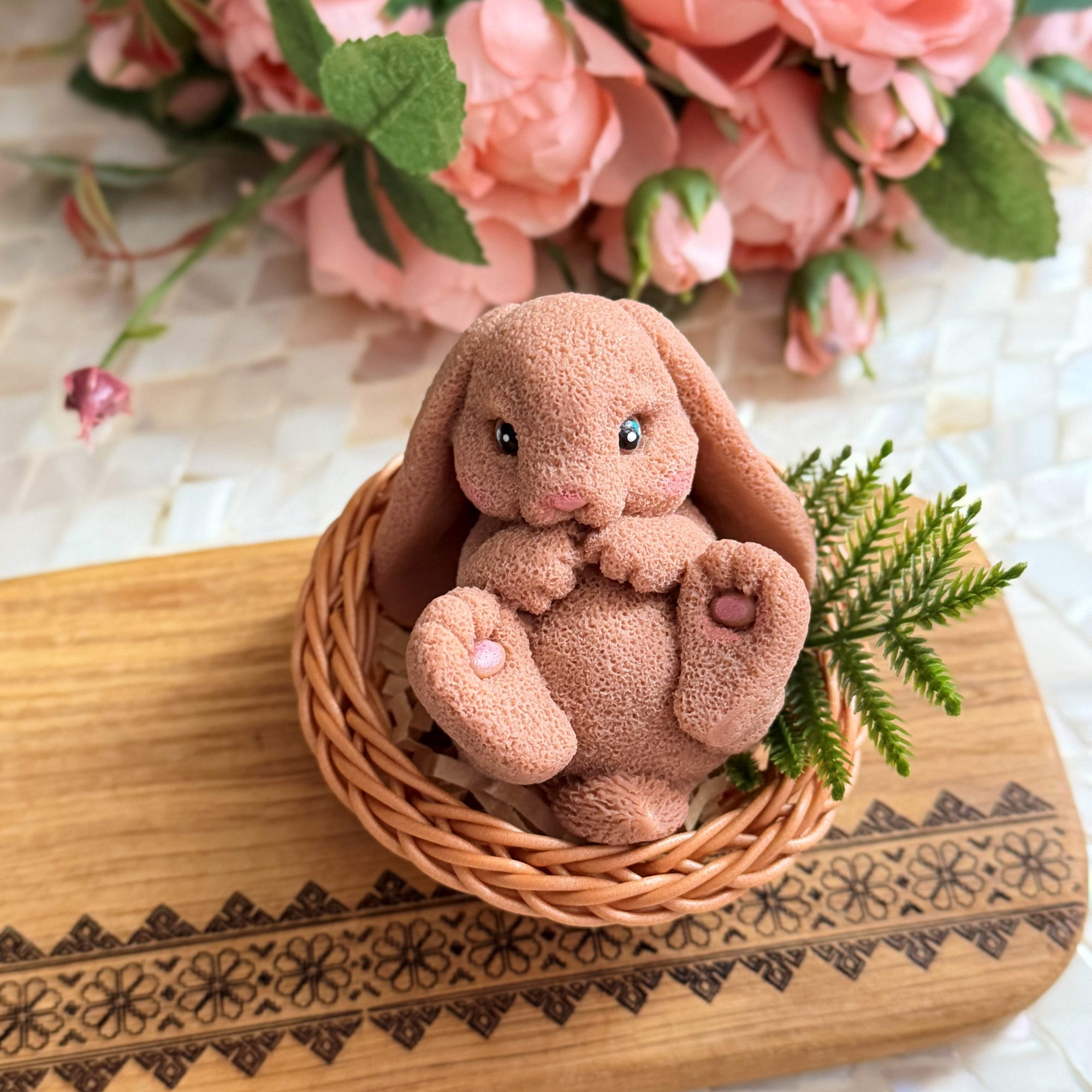 Plush bunny toy in a wicker basket on a decorative wooden board with flowers in the background.