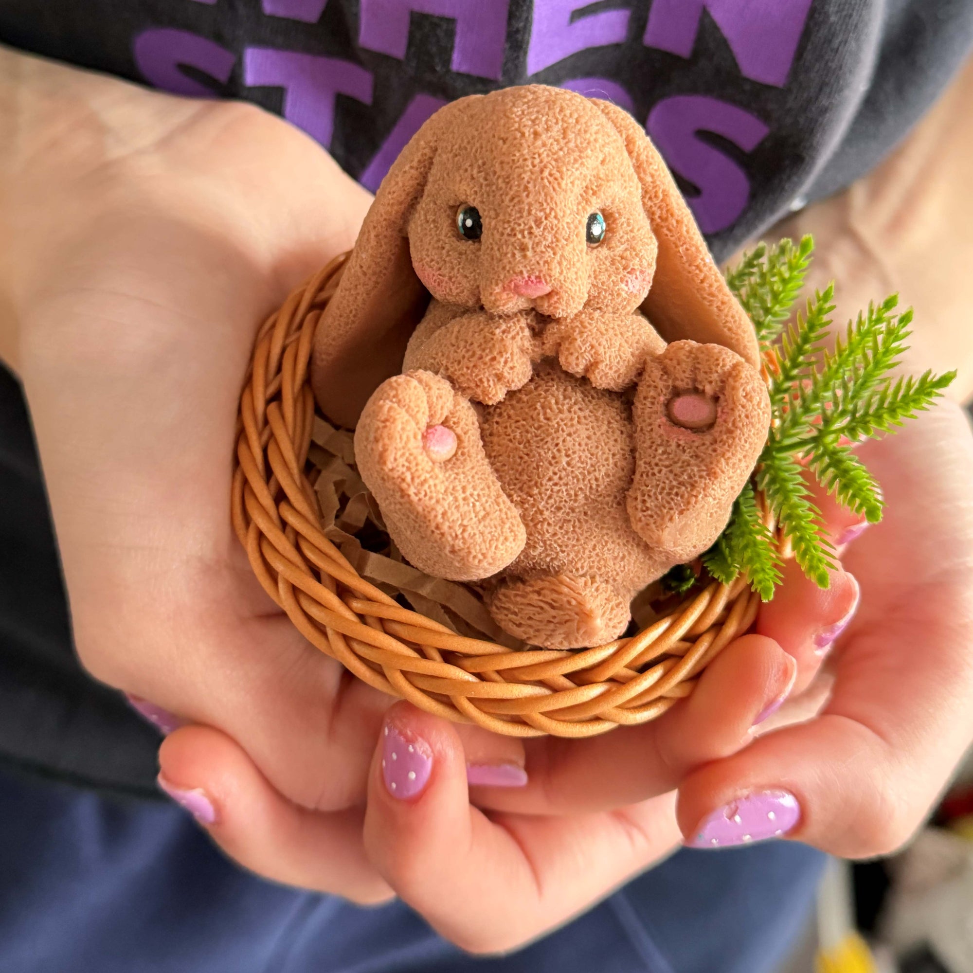 Small brown rabbit plush toy in a wicker basket held by hands with purple nail polish.