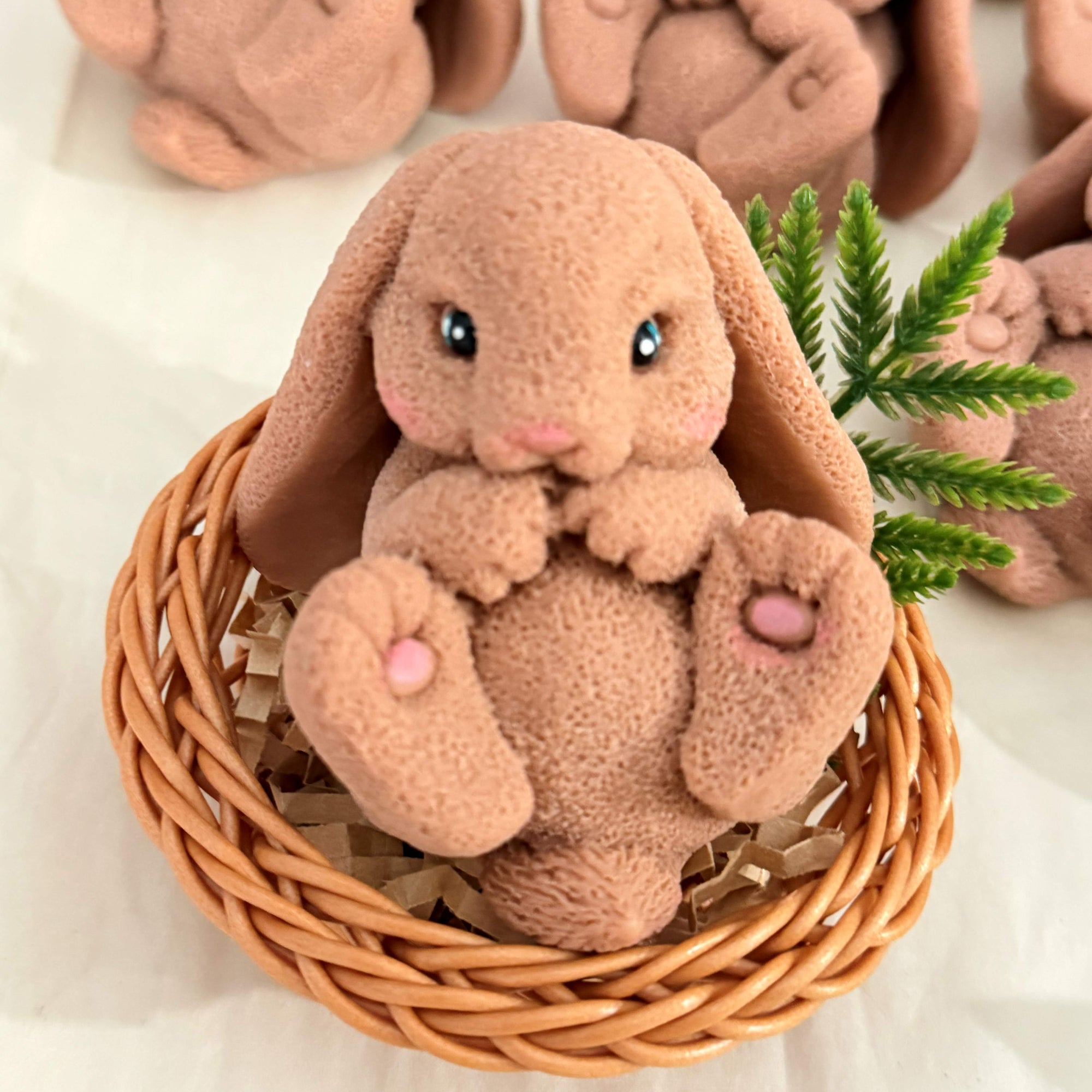 A detailed bunny-shaped soap sitting upright in a small woven-style basket with filler, with similar bunny soaps in the background.