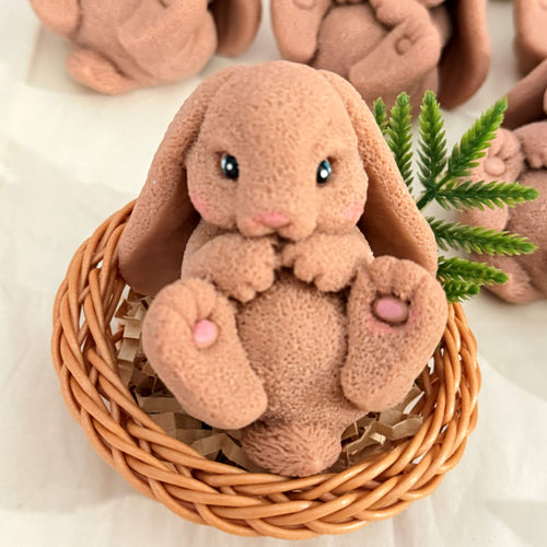 A detailed bunny-shaped soap sitting upright in a small woven-style basket with filler, with similar bunny soaps in the background.