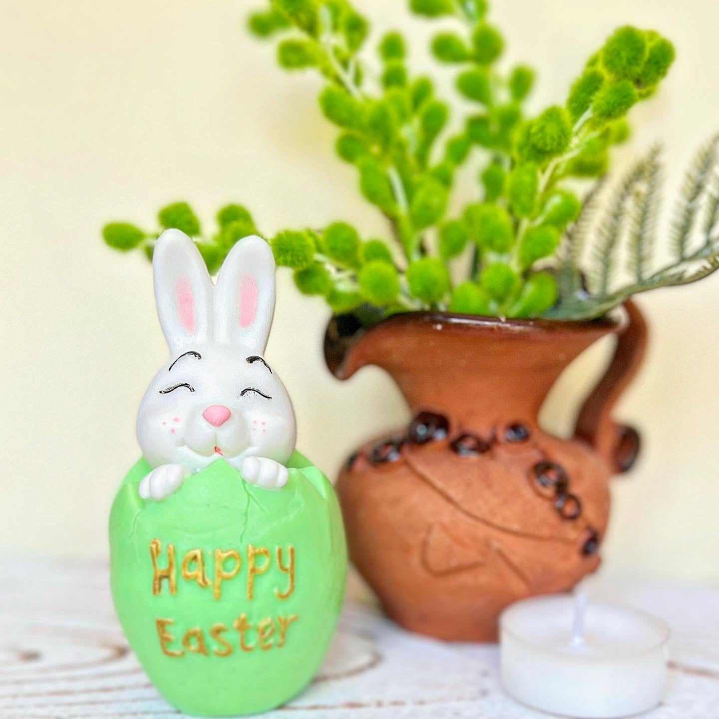 Decorative Easter egg with a bunny on top, featuring 'Happy Easter' text, in front of a brown vase with greenery.
