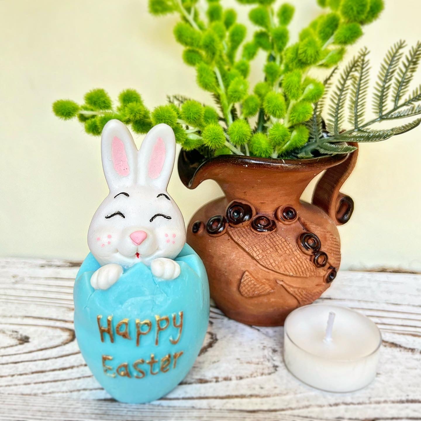 Decorative Easter egg with a bunny face, 'Happy Easter' text, and a wooden vase with plants on a wooden surface.