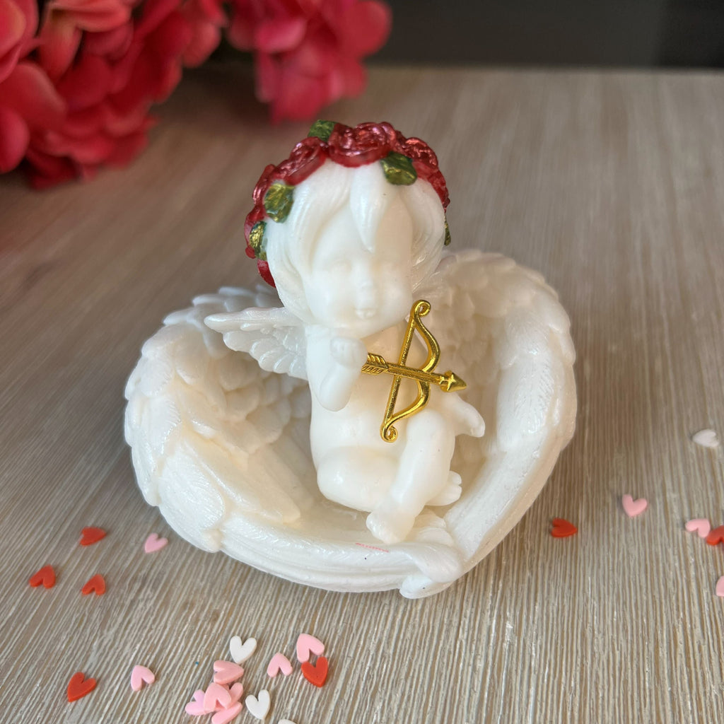 Small angel figurine with a gold bow in a white shell on a wooden surface with red and pink flowers in the background.