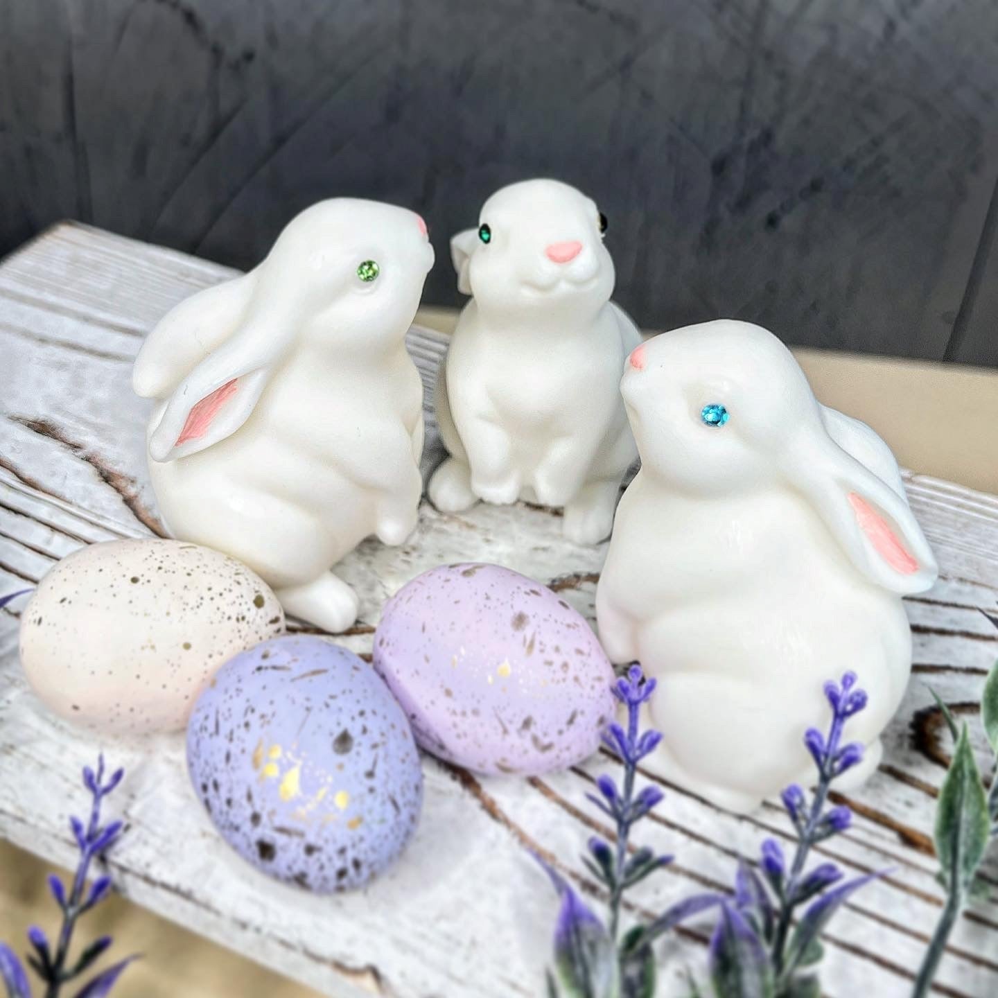 Three white rabbit figurines with colorful eggs on a wooden surface with lavender flowers.