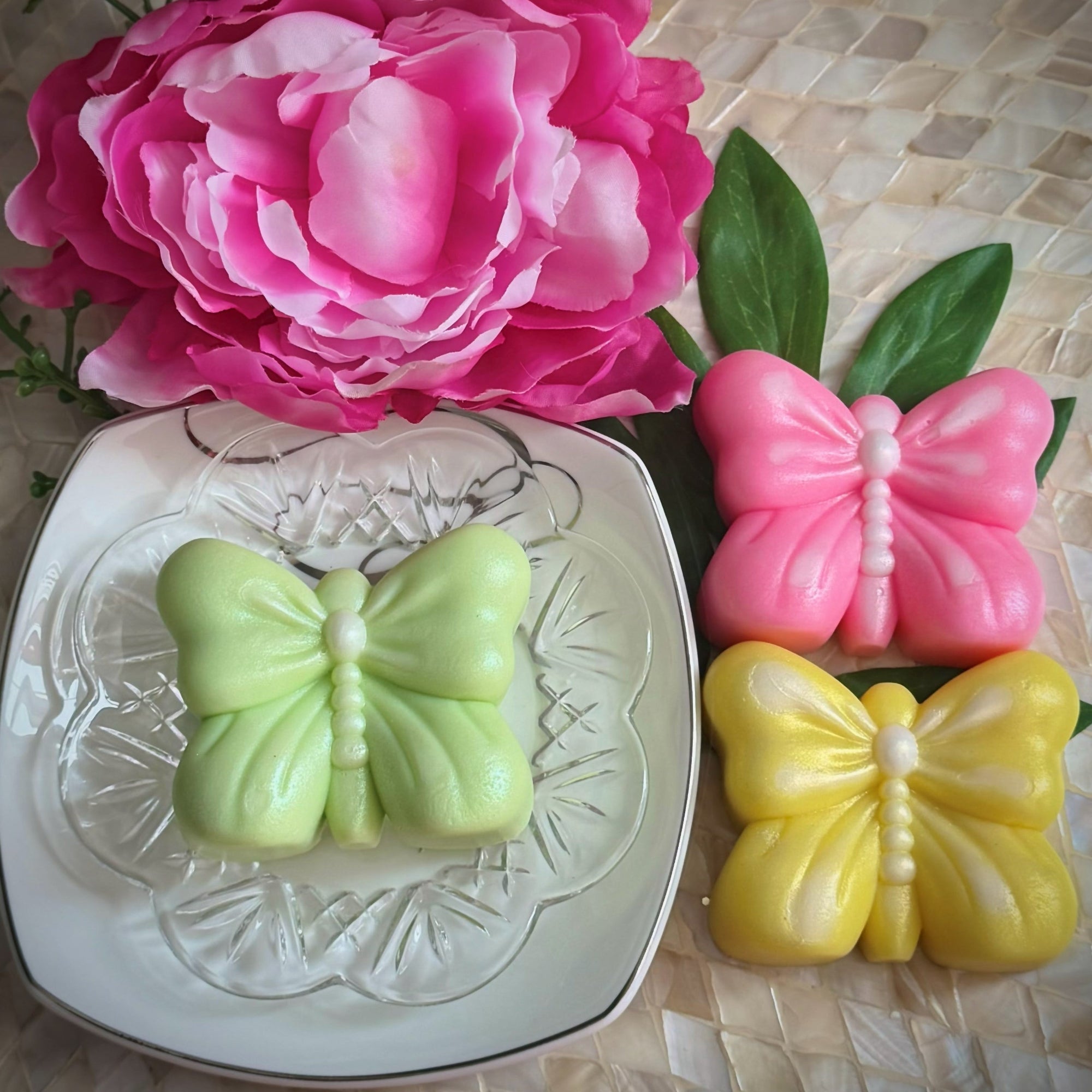Butterfly-shaped soaps in green, pink, and yellow on a textured surface with a large pink flower in the background.