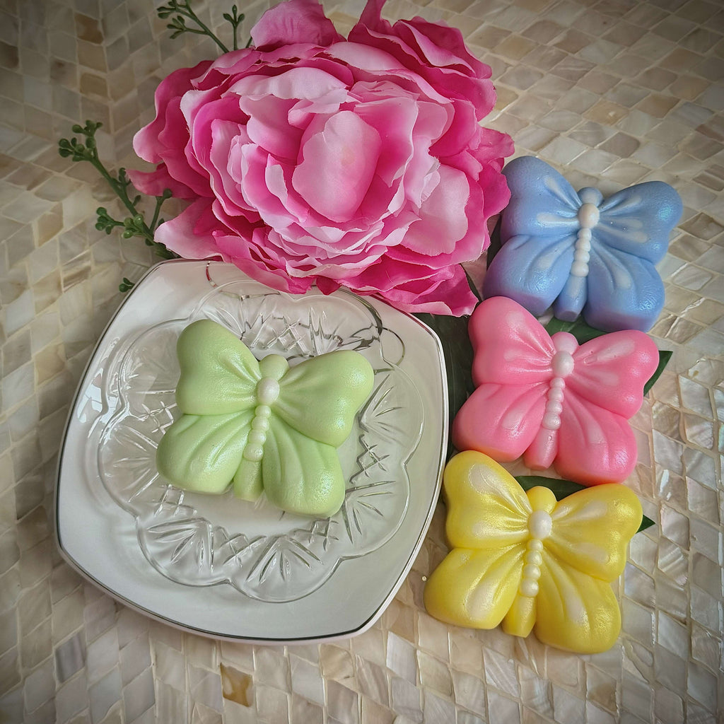 Butterfly-shaped soaps in green, pink, and yellow on a decorative plate with a pink flower in the background.