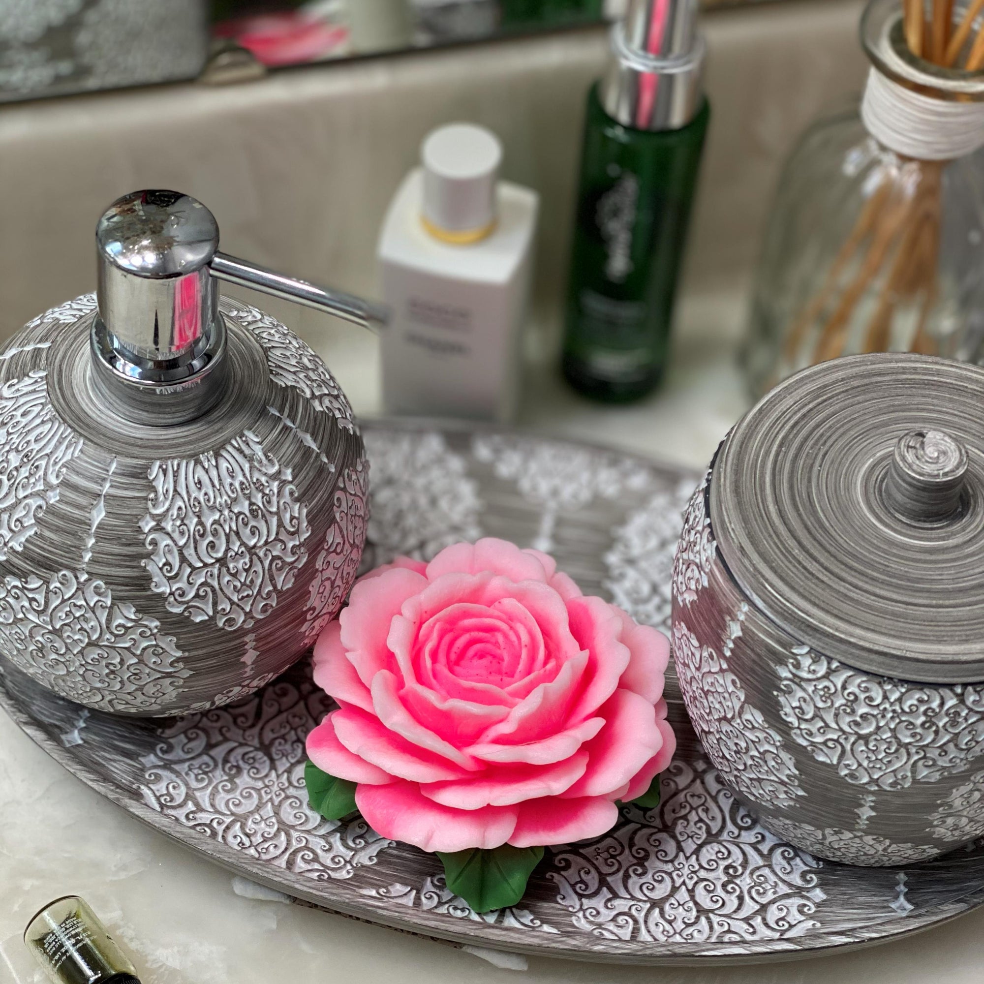 Decorative soap dispenser and candle on a tray with a pink flower in the center, against a blurred background of bottles.