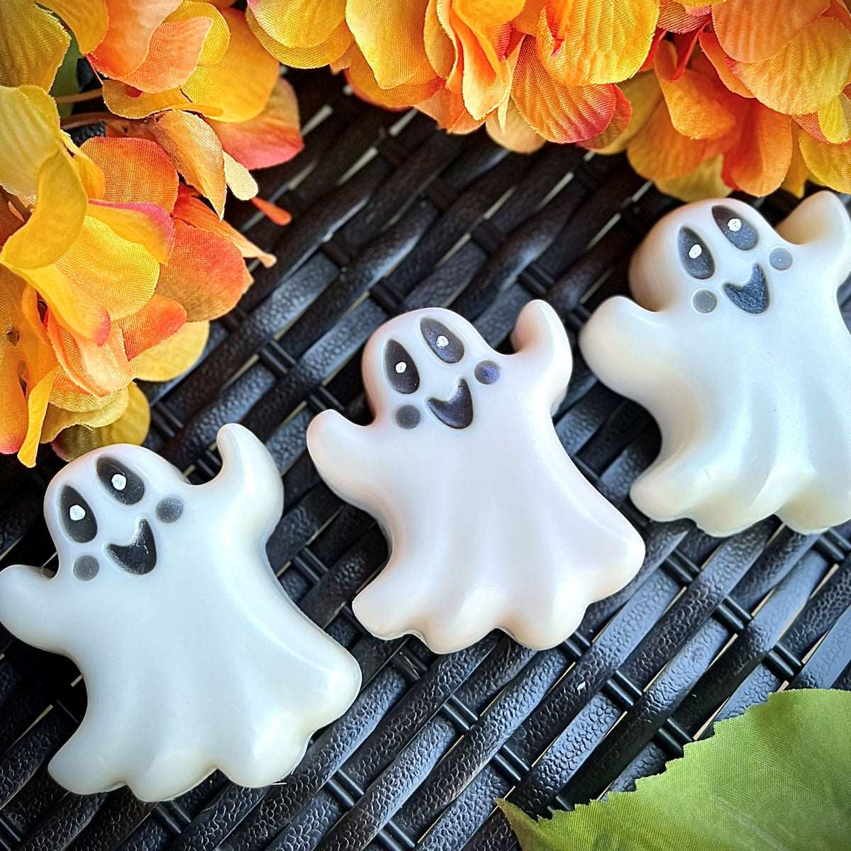 Three white ghost-shaped cookies with black facial features arranged on a black woven surface, with orange and yellow flowers in the background and a green leaf in the corner.