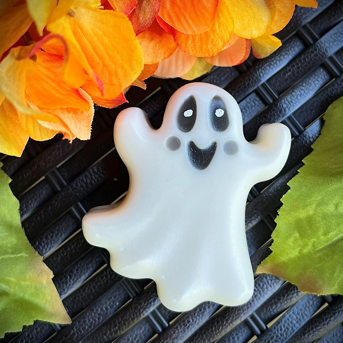 A small ghost-shaped white item with black facial features, photographed on a dark woven surface with orange flowers and green leaves around it.