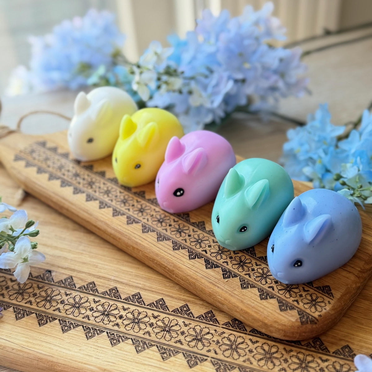 Five colorful rabbit-shaped items on a wooden board with flowers in the background