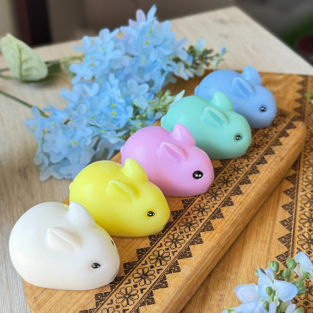 Five small bunny-shaped soaps in white, yellow, pink, light green, and light blue arranged on a patterned wooden tray with blue flowers nearby.